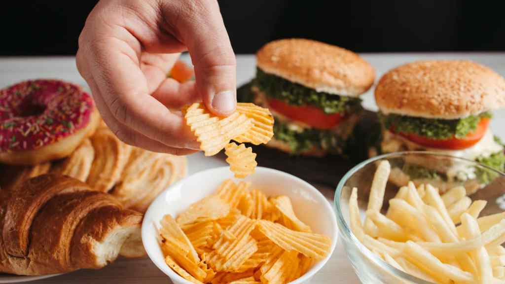 A hand takes a ridged potato chip from a bowl, with other snacks including French fries, burgers, a croissant, and a donut visible on the table.