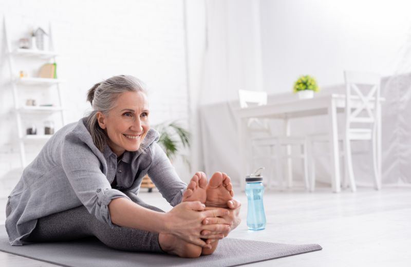 Senior woman smiling and stretching on a yoga mat in a bright, modern living room.
