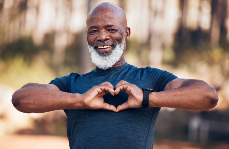 An elderly black man with a white beard smiling and making a heart shape with his hands in a park.