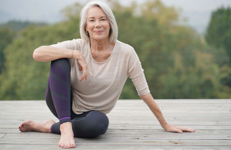 Elderly woman smiling while sitting in a yoga pose outdoors on a wooden deck, with a blurred natural background.
