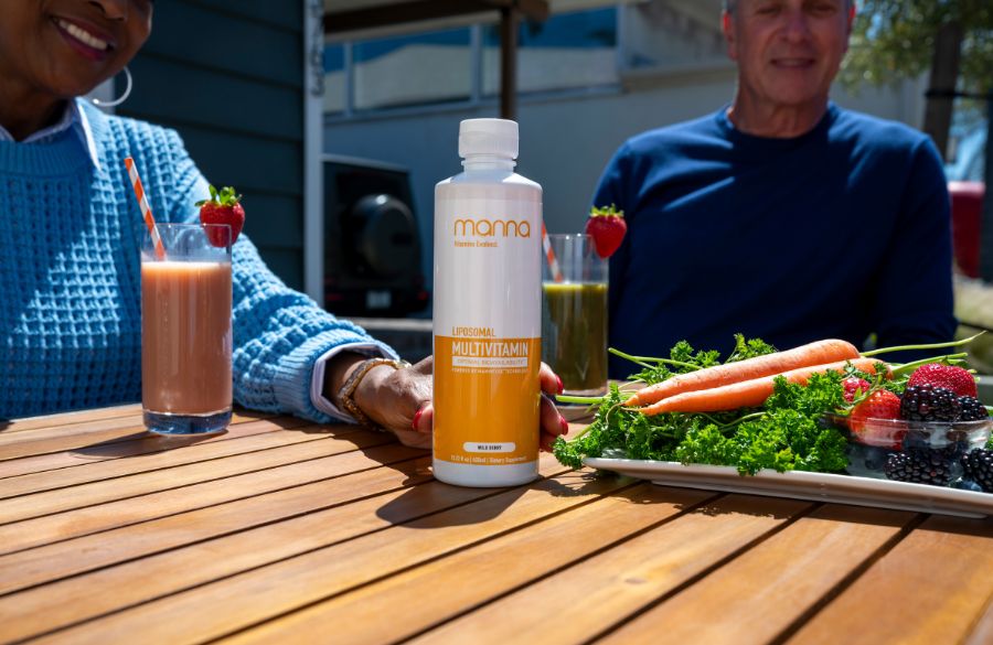 A senior couple sits at a wooden table with healthy foods and a bottle labeled "multivitamin," enjoying fresh smoothies in a sunny outdoor setting.