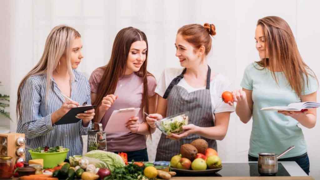 Four women engaged in meal preparation and recipe consultation in a kitchen.