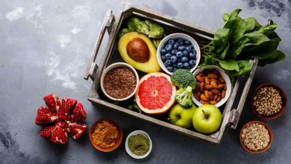 A variety of fresh fruits, vegetables, nuts, and seeds arranged neatly in a wooden crate on a textured gray surface.