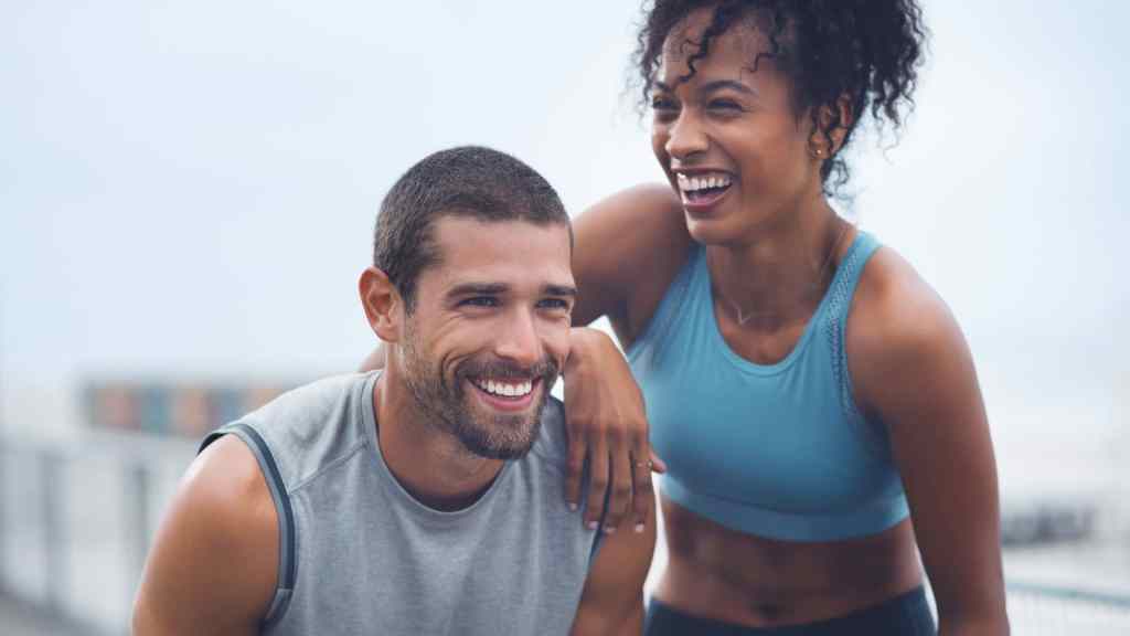A man and woman in athletic attire smiling outdoors, with the woman leaning her arm on the man's shoulder.