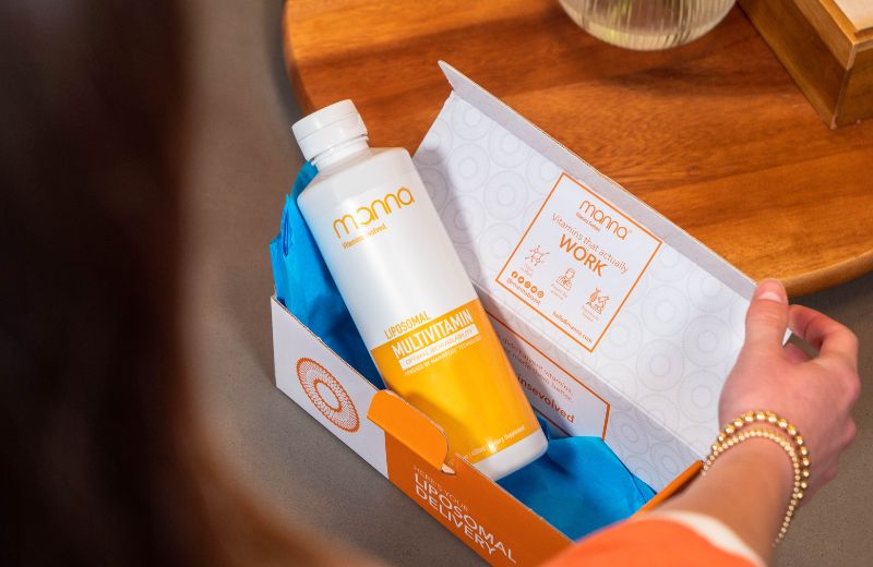 Person opening a box containing a large bottle labeled "Liposomal Multivitamin" from a brand named "Manna." The box interior includes text and icons related to the product.