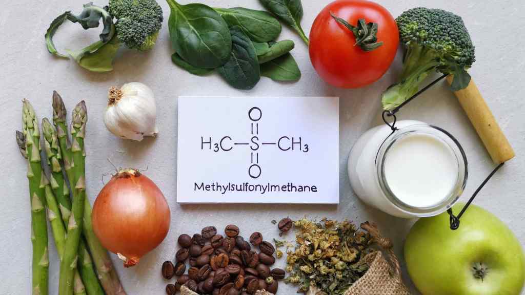 Various foods like asparagus, onion, spinach, tomato, and broccoli surround a card showing the chemical structure of Methylsulfonylmethane, alongside a jar of milk and coffee beans.