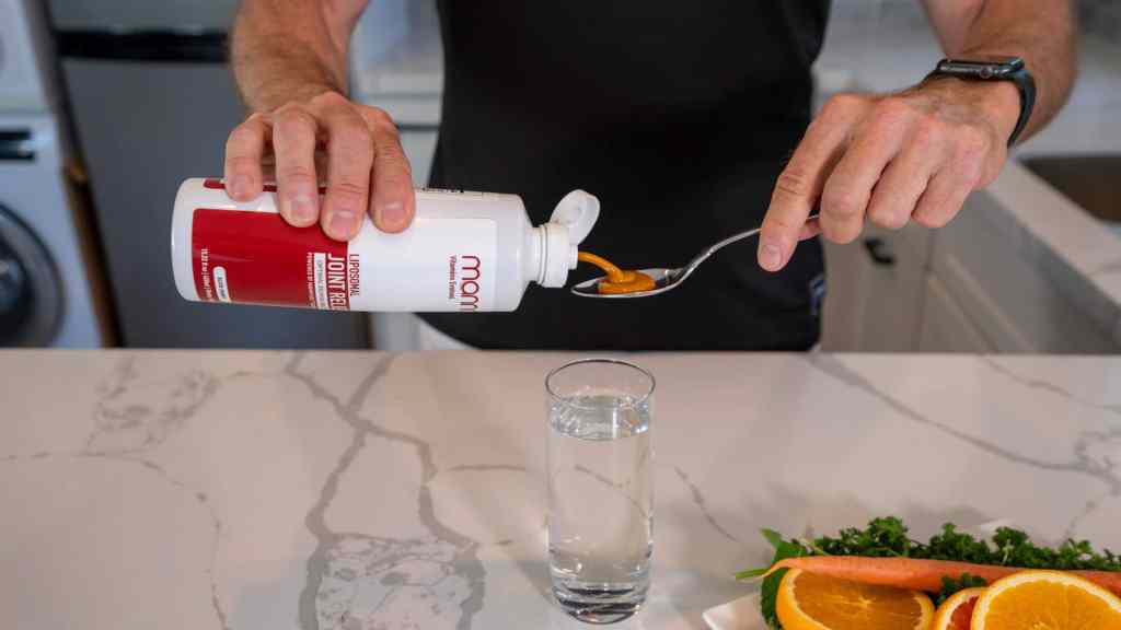 A person pours liquid from a bottle into a spoon over a glass of water on a kitchen counter. Slices of orange, carrot, and greens are in view.