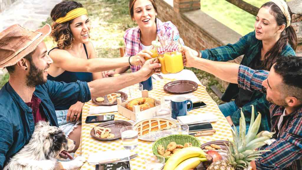 Group of friends toasting with juice glasses at an outdoor picnic table. Various fruits and pastries are on the table, along with a dog sitting by the side.