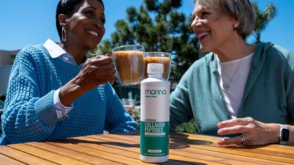Two women clinking glasses while sitting outdoors at a table with a bottle of Liposomal Collagen Peptides by Mama Protein Coated placed in the foreground.