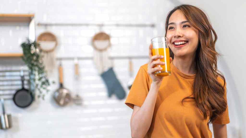 Woman in a kitchen holding a glass of orange juice, smiling, with cooking utensils in the background.