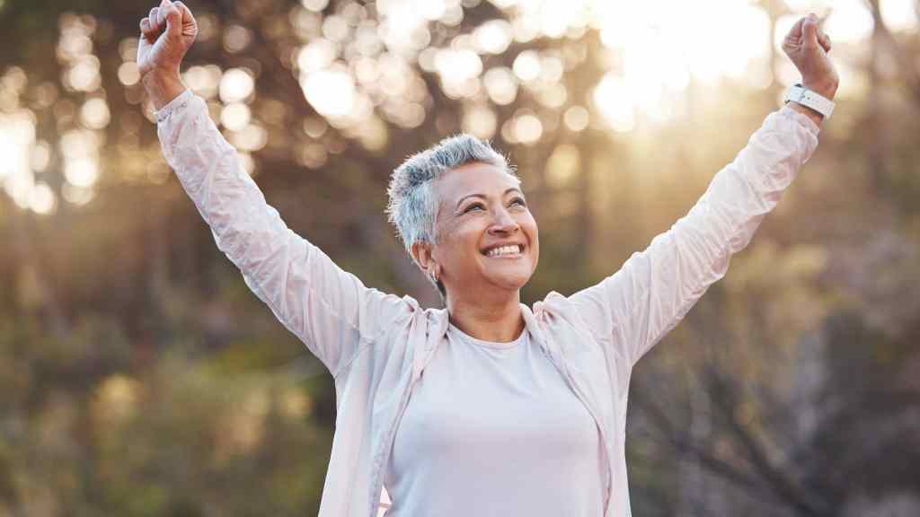 A person with short gray hair is smiling and raising their arms in triumph outdoors with a blurred forest background.