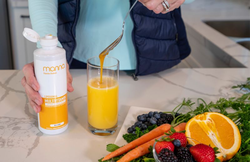 A person stirring a glass of orange juice with a fork next to an open bottle of liposomal multivitamin, surrounded by fruits and vegetables.