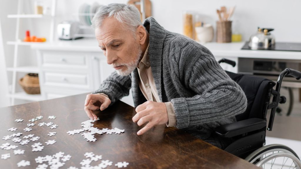 Elderly man in a wheelchair working on a puzzle at a wooden table in a bright kitchen.