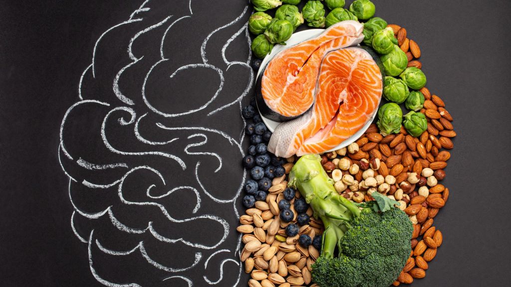 A chalk-drawn brain on a blackboard is paired with salmon, nuts, broccoli, blueberries, Brussels sprouts, and other healthy foods arranged beside it.
