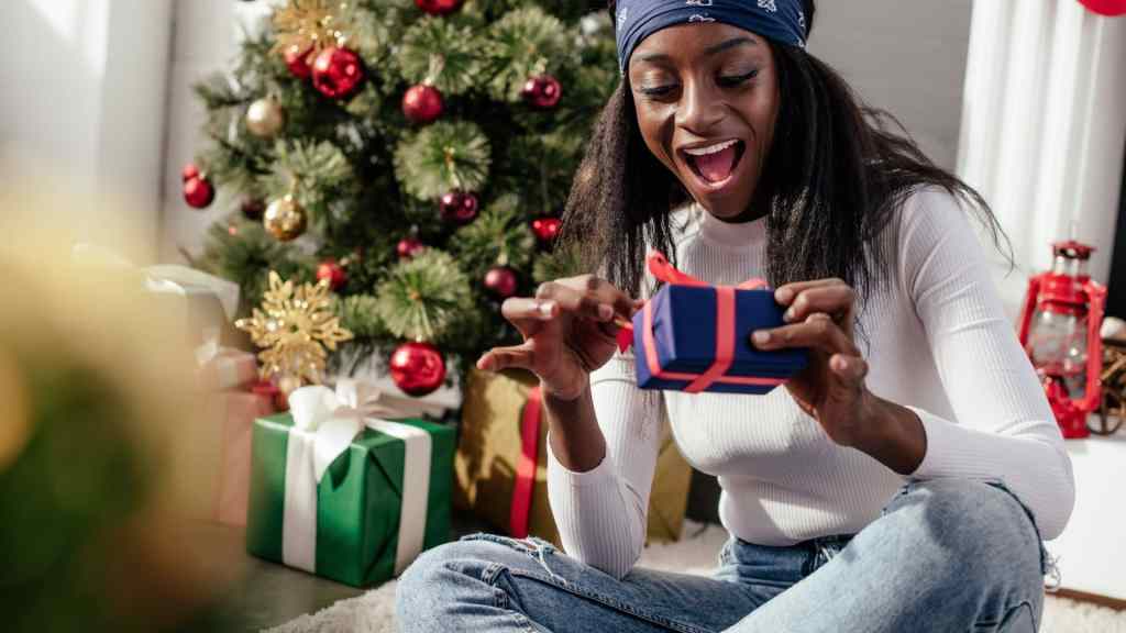 A woman sits on the floor, excitedly opening a gift near a decorated Christmas tree.