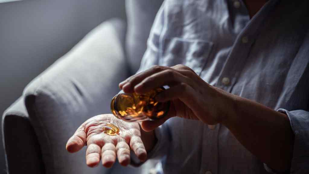 A person pours yellow capsules from a brown bottle into their hand while sitting on a couch.