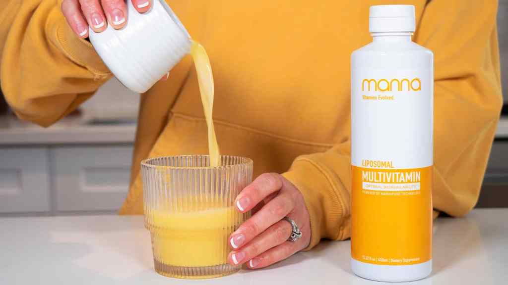 A person in a yellow sweatshirt pours a yellow liquid from a small container into a glass next to a bottle labeled “Manna Liposomal Multivitamin.”.