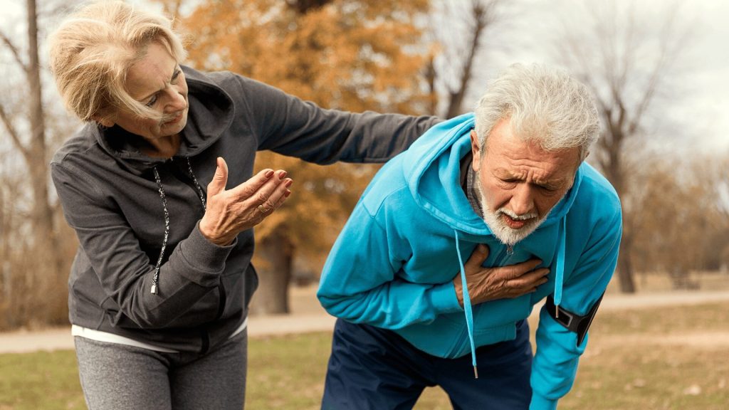 An elderly man in a blue hoodie clutches his chest in pain while a concerned elderly woman in a gray hoodie supports him outdoors on a cloudy day.