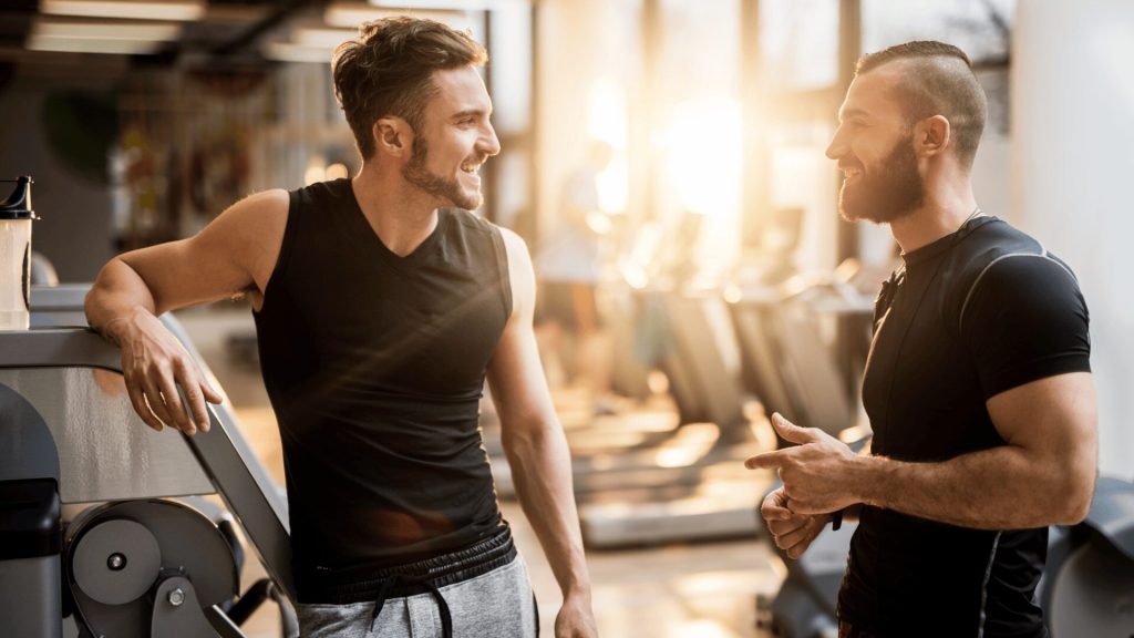 Two men in athletic wear are smiling and talking in a gym filled with exercise equipment, backlit by sunlight from large windows.