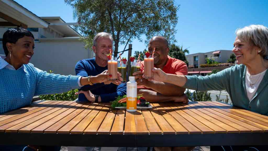 Four people sitting at an outdoor table, holding up glasses with drinks, smiling. A sunscreen bottle is on the table.