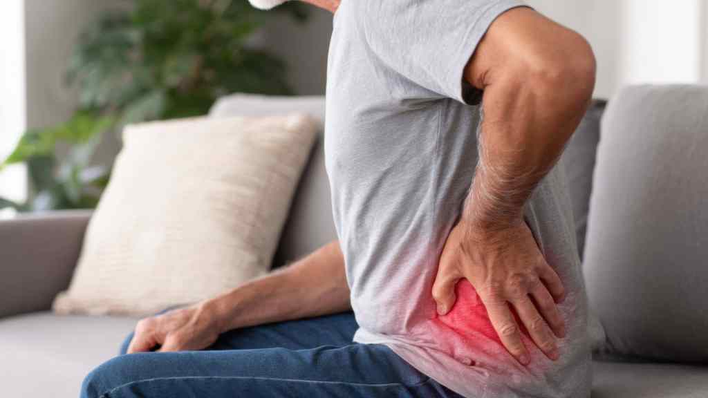 A person sitting on a couch holds their lower back in discomfort, with a red highlight indicating pain in the area.
