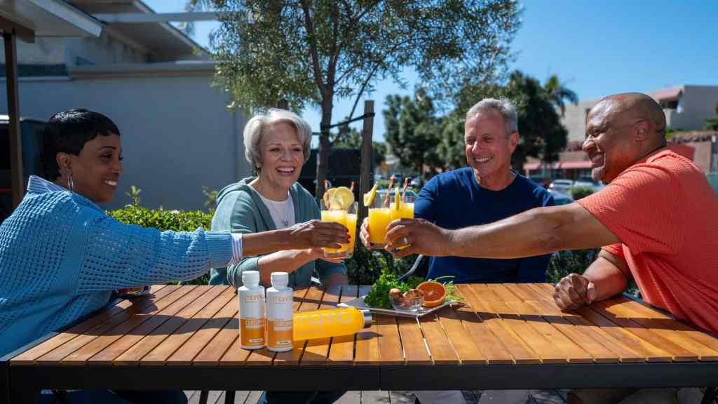 Four adults sit outside at a wooden table, smiling and clinking glasses of orange juice, with bottles and fruit on the table.