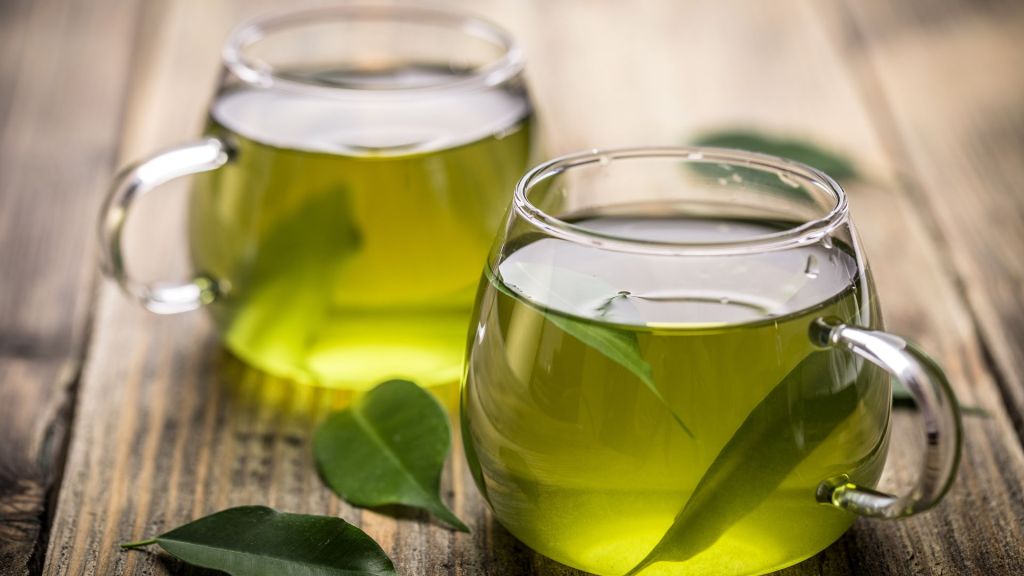 Two glass cups of green tea with green leaves inside, placed on a wooden surface with additional leaves scattered around.