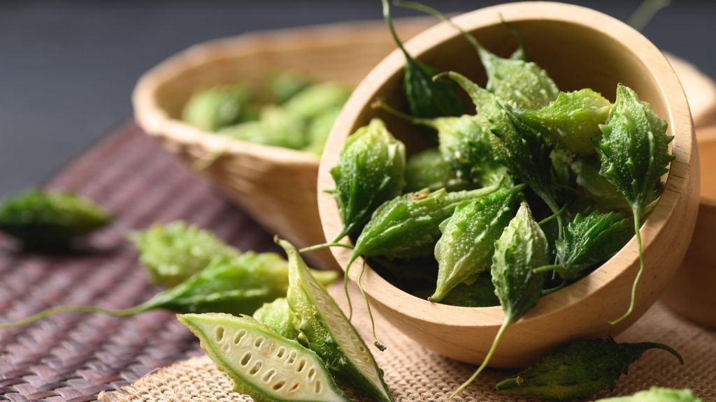 Several fresh green bitter melons, some whole and some sliced, are arranged in wooden bowls on a woven mat.