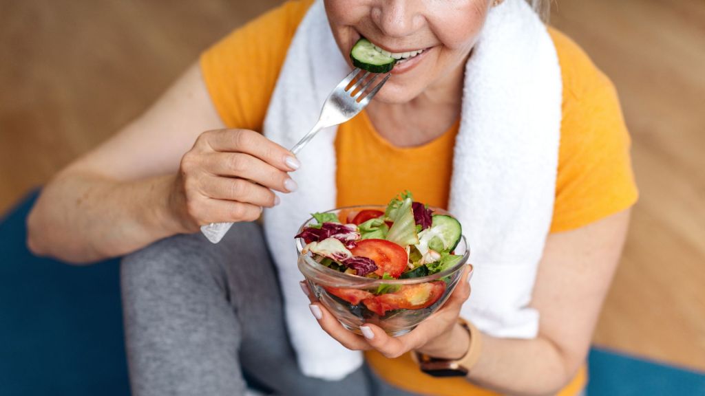 A person wearing a yellow shirt and white towel eats a salad with a fork while sitting on a blue mat.