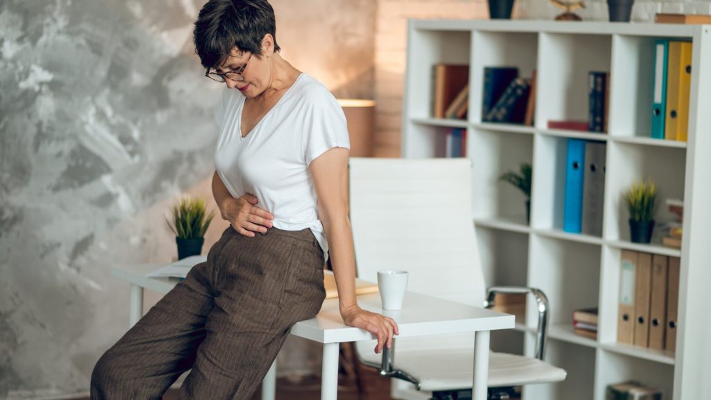 A person with short hair and glasses, wearing a white T-shirt and brown pants, leans against a desk, holding their stomach, in what appears to be an office setting.