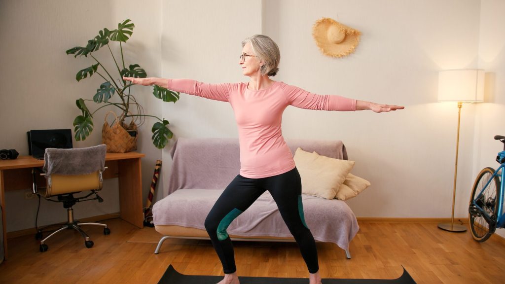 An elderly woman wearing glasses and workout clothes performs a yoga pose on a mat in a well-lit living room with a couch, plants, a desk, and a bicycle.