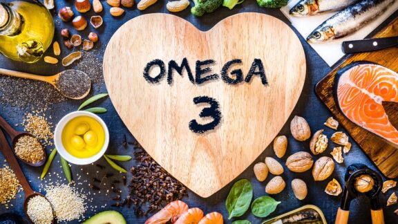 A heart-shaped wooden board with the text "Omega 3" surrounded by foods rich in Omega-3 fatty acids, such as salmon, walnuts, seeds, avocado, and fish.