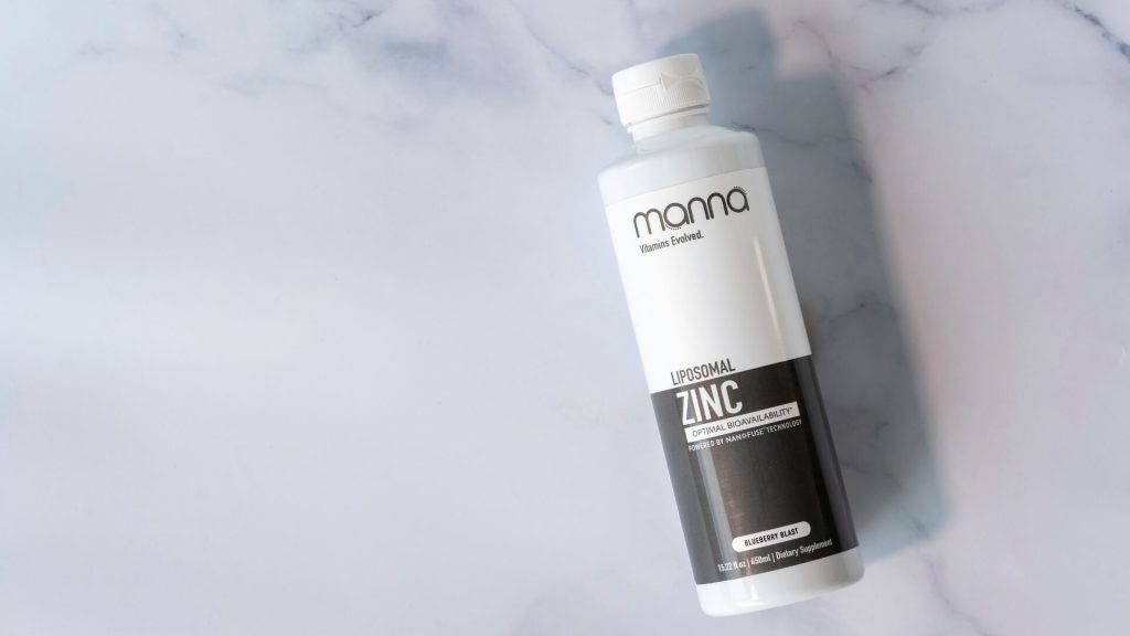 A bottle of Manna Liposomal Zinc supplement on a marble surface. The white bottle has a black label near the bottom with product details.