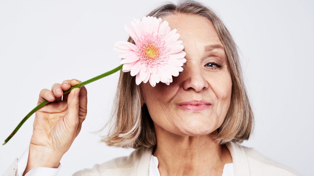 A woman with shoulder-length hair holds a pink flower over one eye and smiles gently against a plain background.