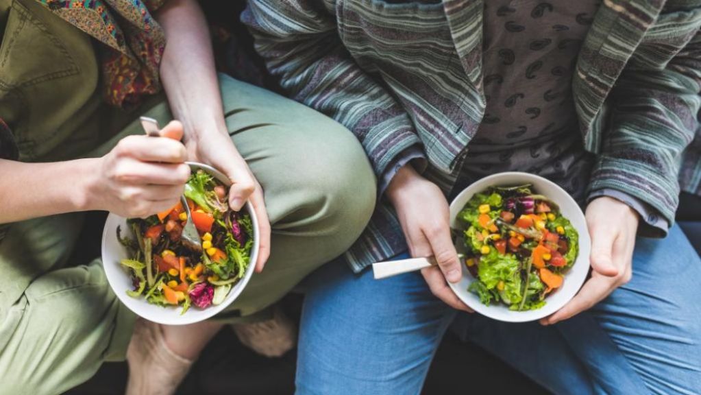 Two individuals sitting close together holding bowls of colorful salad.