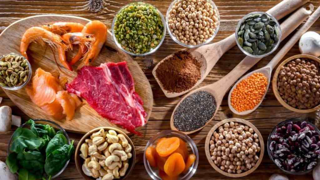 A variety of proteins and legumes displayed on wooden surface, including seafood, red meat, nuts, and seeds.