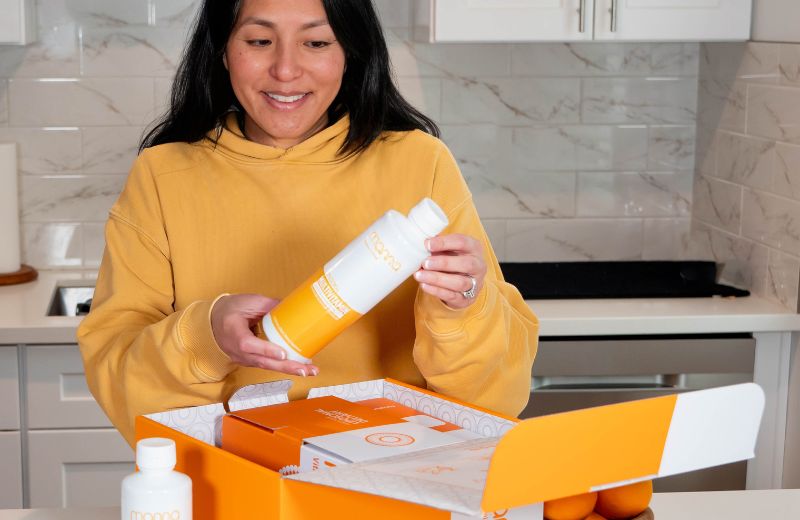 Woman in a yellow sweater unpacking a bottle from a subscription box in a modern kitchen.