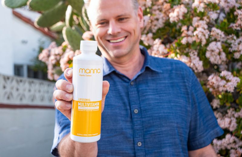 A person in a blue shirt holds a bottle of Manna Liposomal Multivitamin in an outdoor setting with blooming flowers in the background.