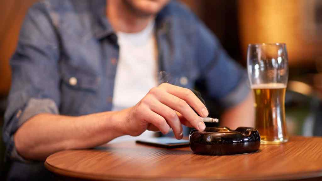 Person holding a cigarette above an ashtray on a table beside a pint of beer and a smartphone.