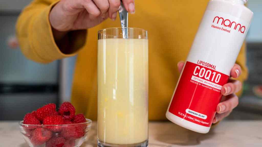 Person in yellow sweater stirs a glass of yellow liquid with a spoon, holding a bottle of Manna Liposomal CoQ10. A bowl of raspberries is nearby on the countertop.