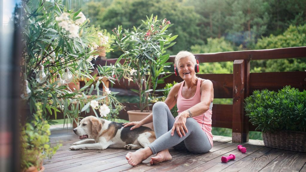 An elderly woman wearing headphones sits on a wooden deck with a dog beside her. She is dressed in workout clothes, and there are dumbbells on the floor. The area is surrounded by plants and trees.