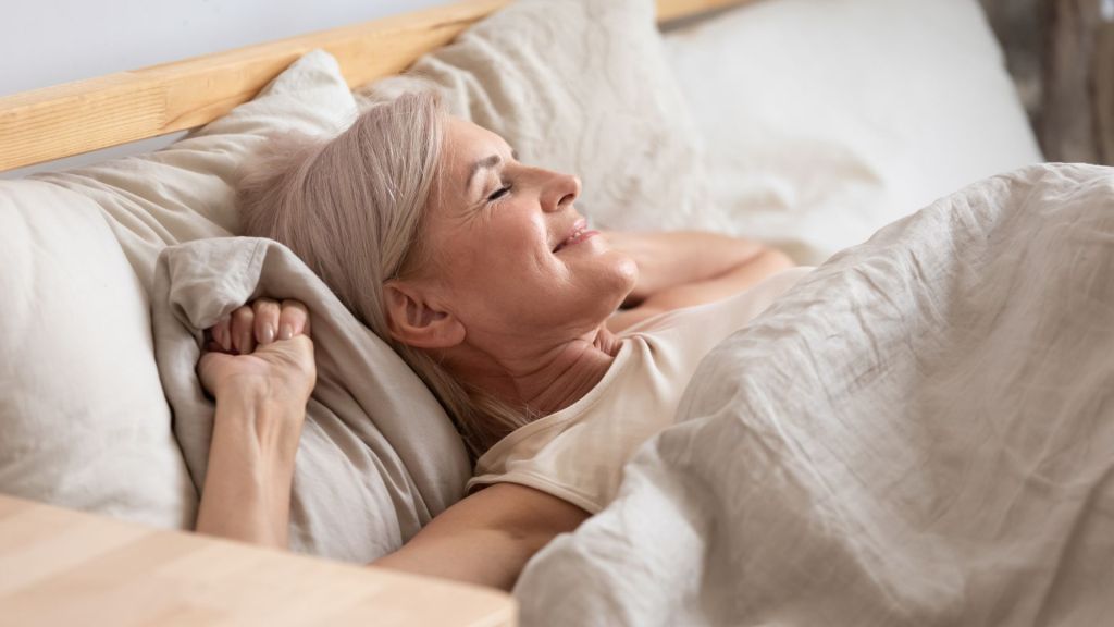An older woman with light hair lies in bed on white linens, stretching and smiling as she wakes up.