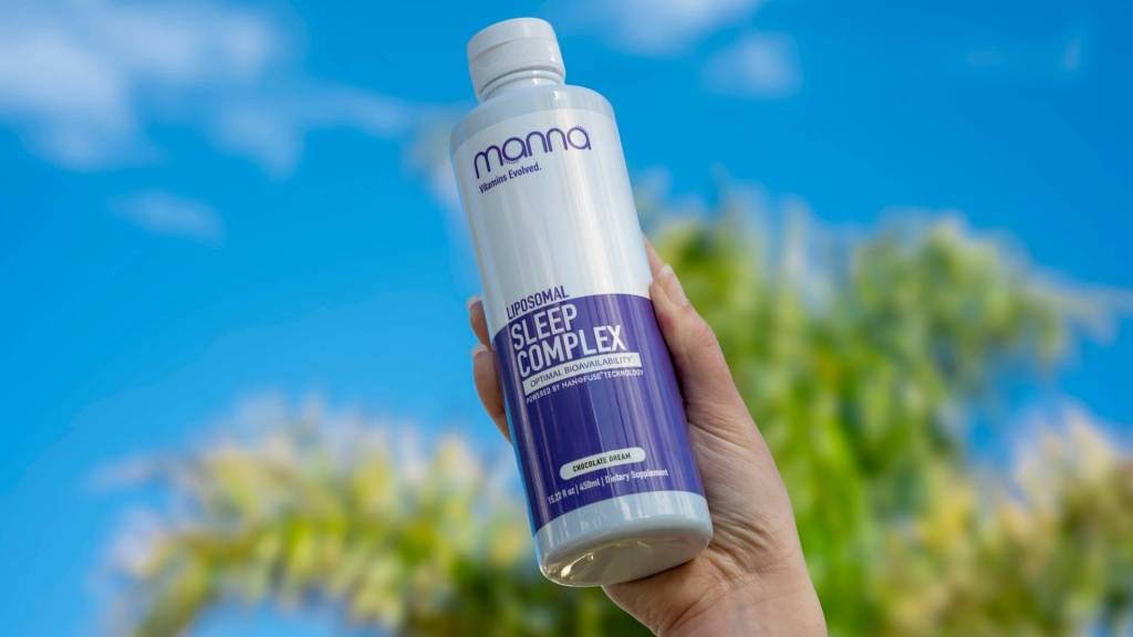 A hand holds a bottle of "manna Liposomal Sleep Complex" against a blue sky background.