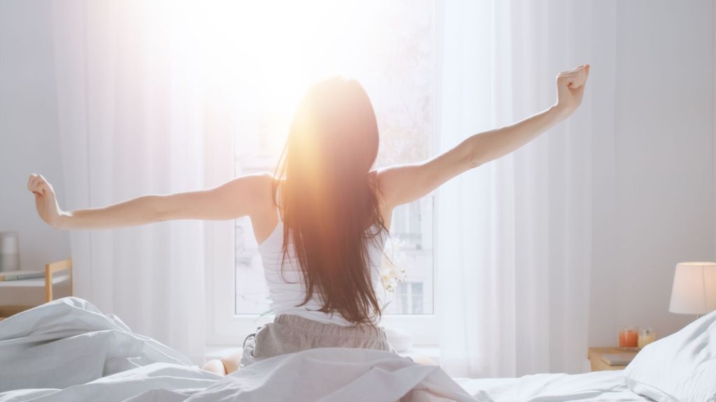 A person sits on a bed stretching with arms outstretched, facing a sunlit window and wearing casual attire.