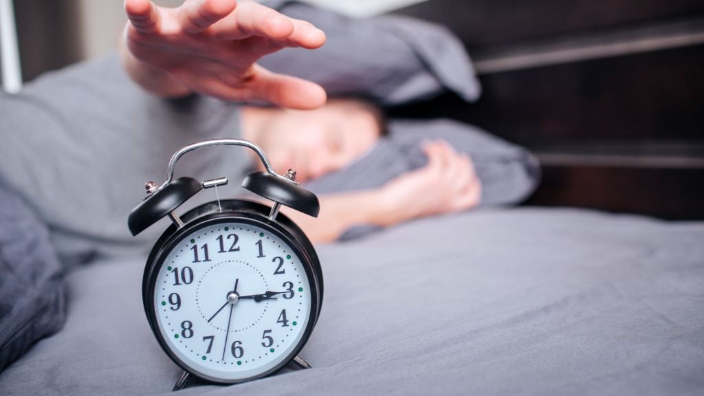 A person reaches for a classic alarm clock on a bed.