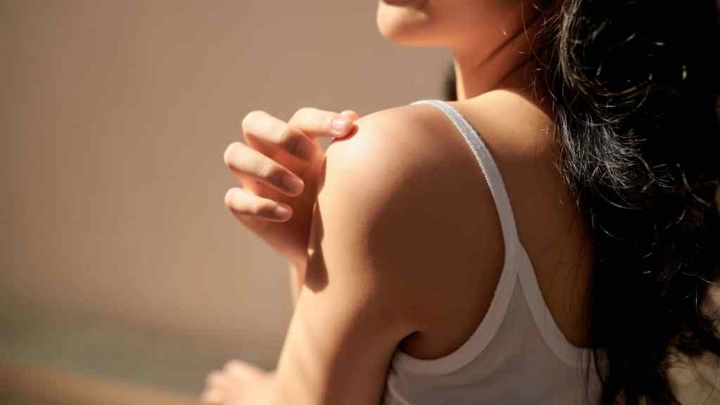 A person in a white tank top gently scratching their shoulder under sunlight.