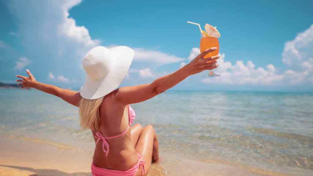 A woman in a pink bikini and white sun hat sits on a beach, holding up an orange drink. The ocean and cloudy sky are in the background.