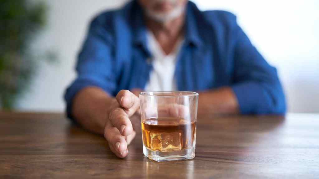 A person sitting at a table, pushing away a glass filled with brown liquid, possibly alcohol, with their hand. The focus is on the glass and hand.