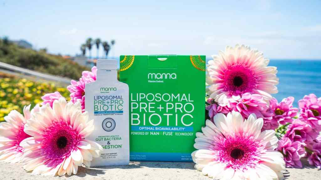 Mama brand liposomal pre+probiotic supplement bottle and box surrounded by pink and white flowers, set outdoors with the ocean and palm trees in the background.
