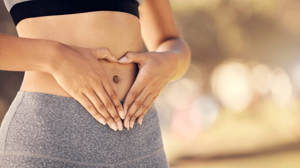 Person wearing workout clothes forms a heart shape with their hands over their bare stomach, outdoors in natural light.